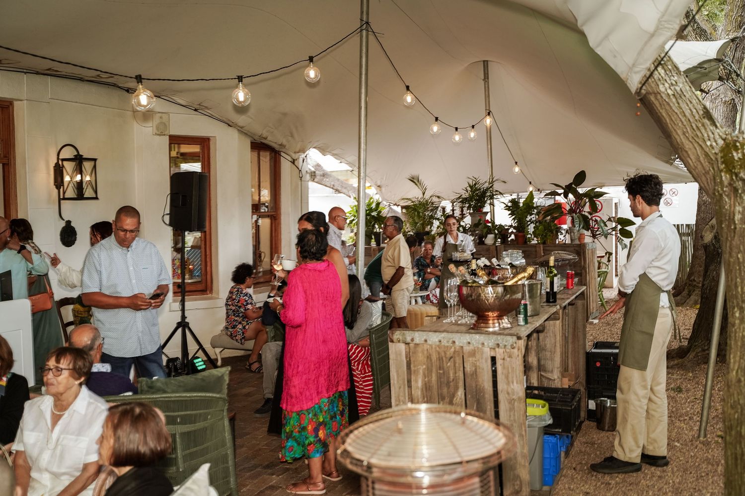 Guests mingling at a lively outdoor garden party under a tent with string lights and food stations.