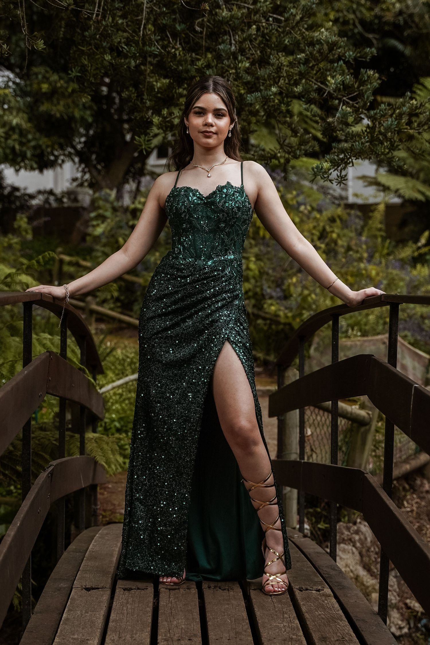 Young woman in emerald green sequin spaghetti strap gown with thigh slit stands on wooden bridge.