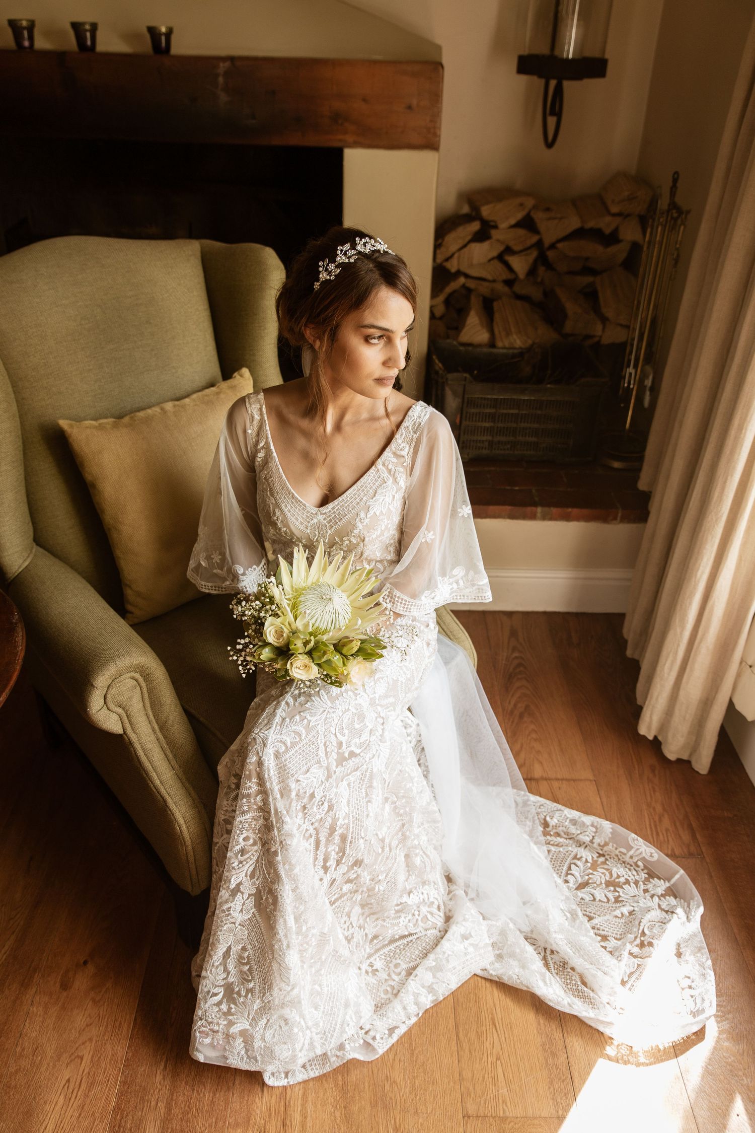 Bride in lace wedding dress with floral crown sitting in armchair holding bouquet.