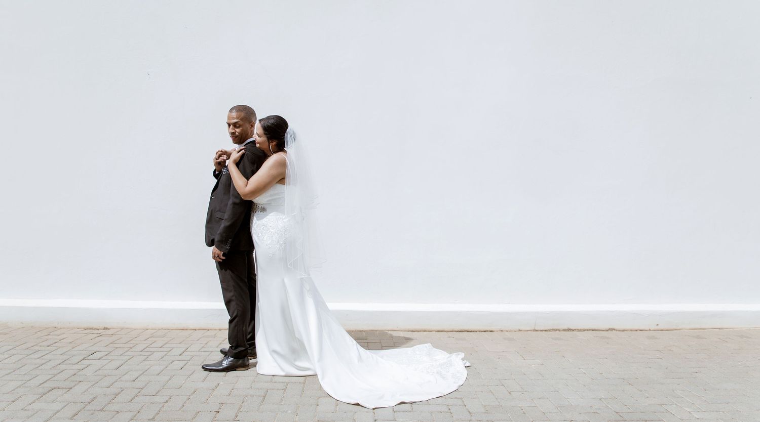 A bride in a white gown and groom in a black suit embrace tenderly against a white wall on their wedding day.