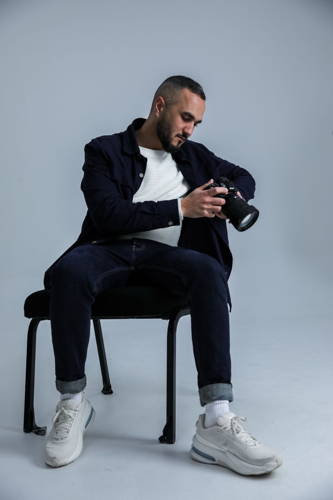 A bearded male photographer sits on a chair reviewing photos on his DSLR camera, wearing dark jacket and jeans.