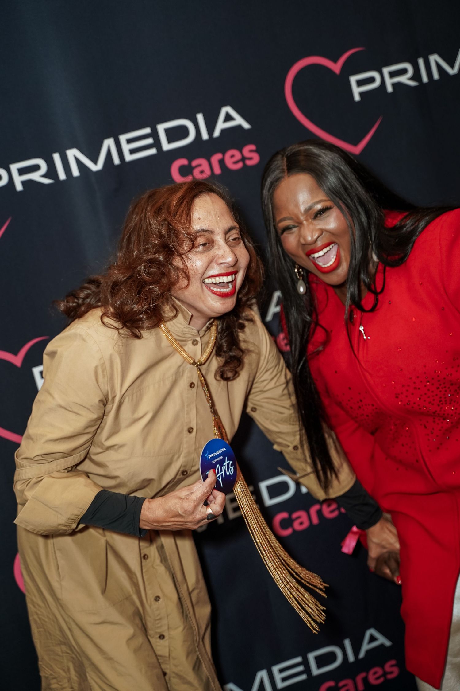 Two women smiling and posing together at a Primedia Cares event backdrop.