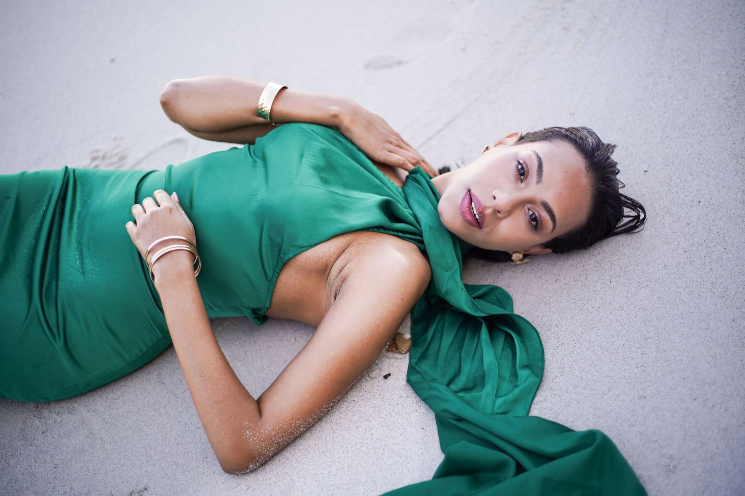 Woman in elegant green satin dress lying gracefully on white sand beach.