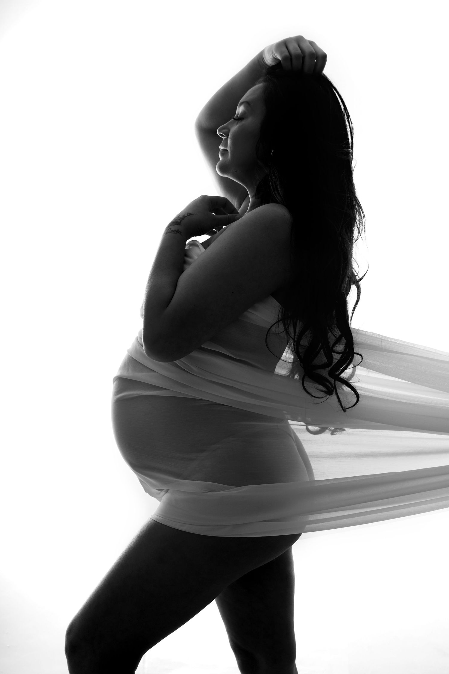 Black and white artistic silhouette of a pregnant woman with long dark hair holding sheer white fabric in motion.
