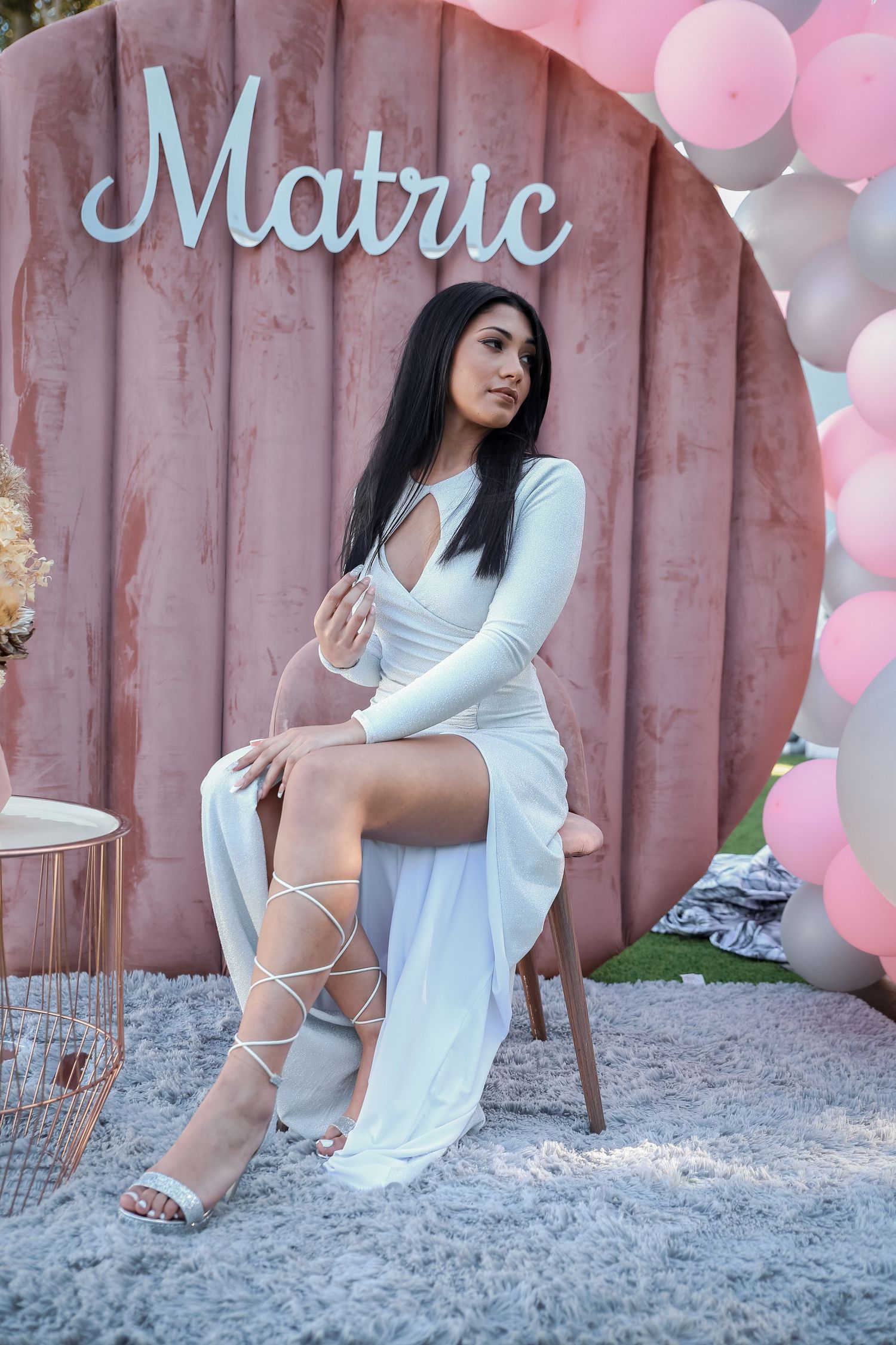 Elegant woman in white long-sleeve cutout dress seated at Matric event with pink balloon decor.
