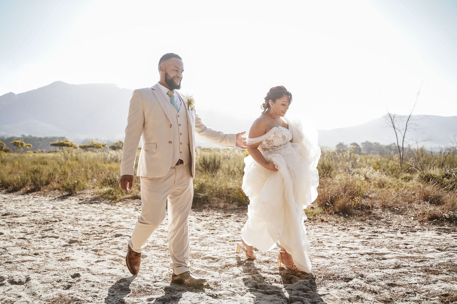 Bride and bearded groom in tan suit walking through sun-drenched arid landscape with mountains.