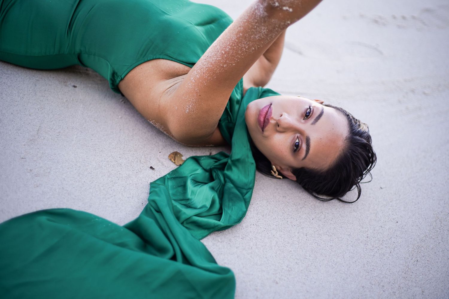 Woman in green satin dress lying on white sand beach looking up at camera.