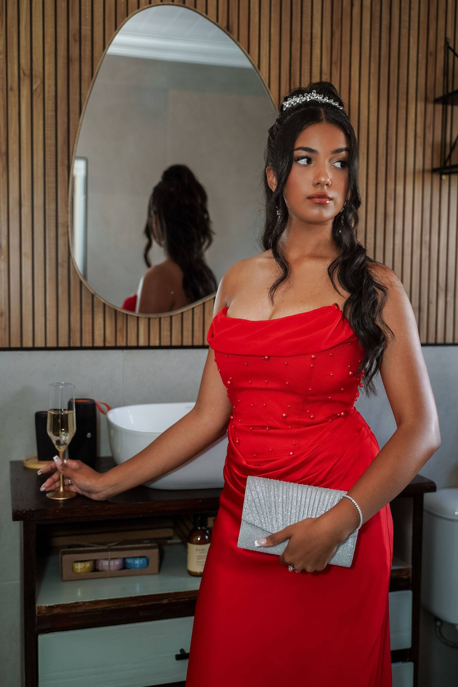 Young woman in red off-shoulder gown with tiara holds champagne flute and clutch near bathroom mirror.
