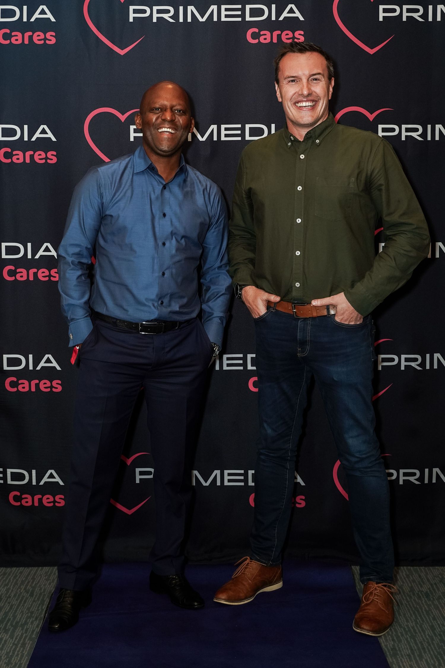 Two men smiling and posing in front of Primedia Cares branded step-and-repeat backdrop.