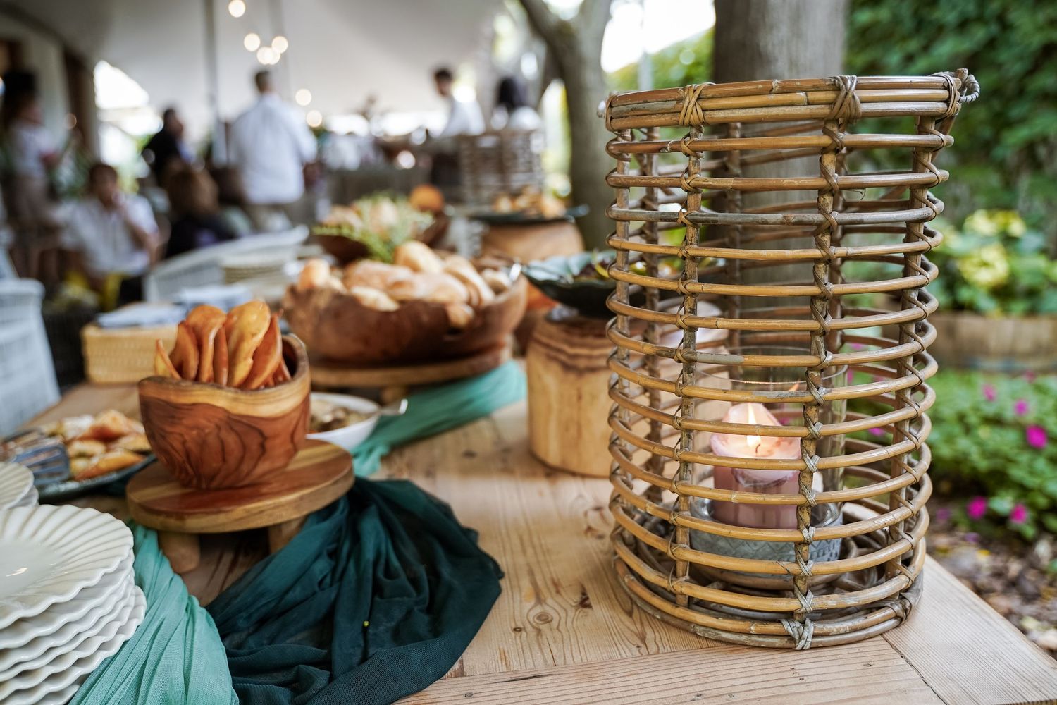 Rustic wooden table with wicker lantern, bread bowls, and food spread at an outdoor garden event.