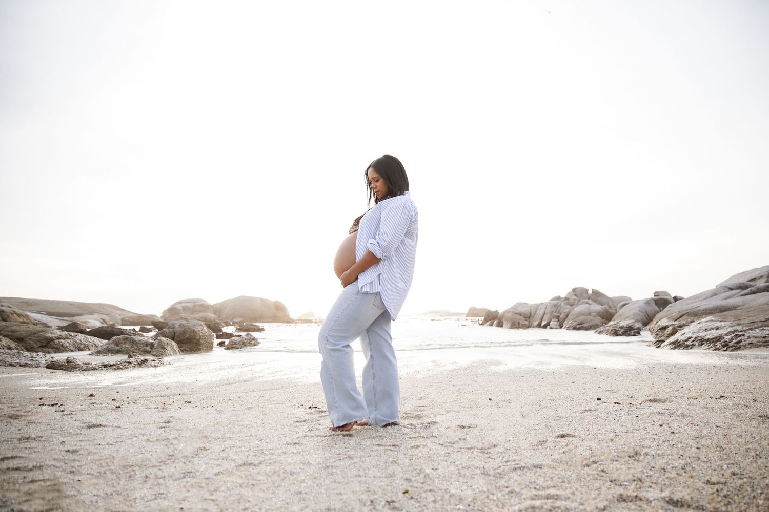 Pregnant woman in white linen outfit standing on sandy beach with rocky coastline backdrop.