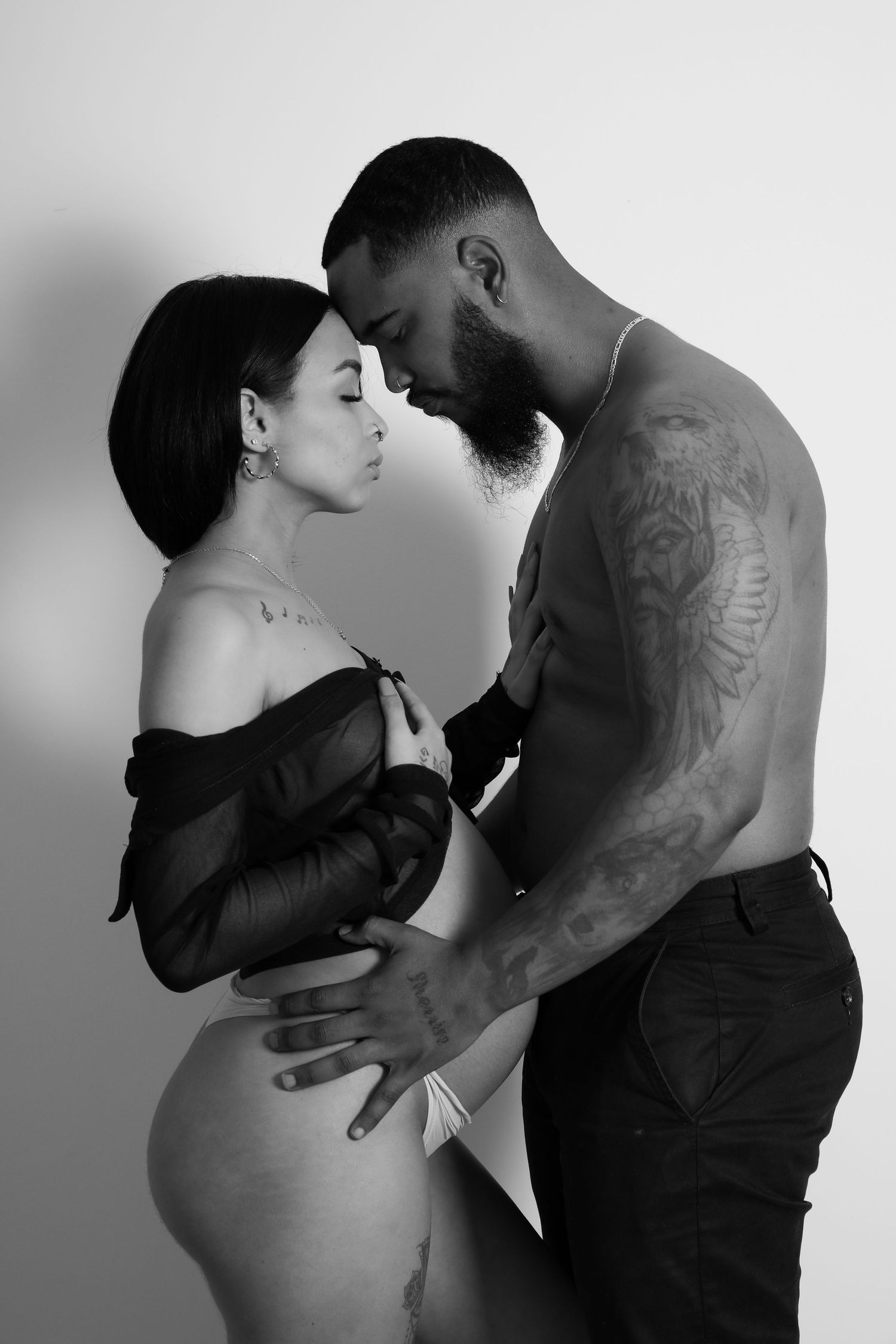 Black and white intimate maternity photo of a tattooed couple embracing, showcasing the pregnant woman's bare belly.