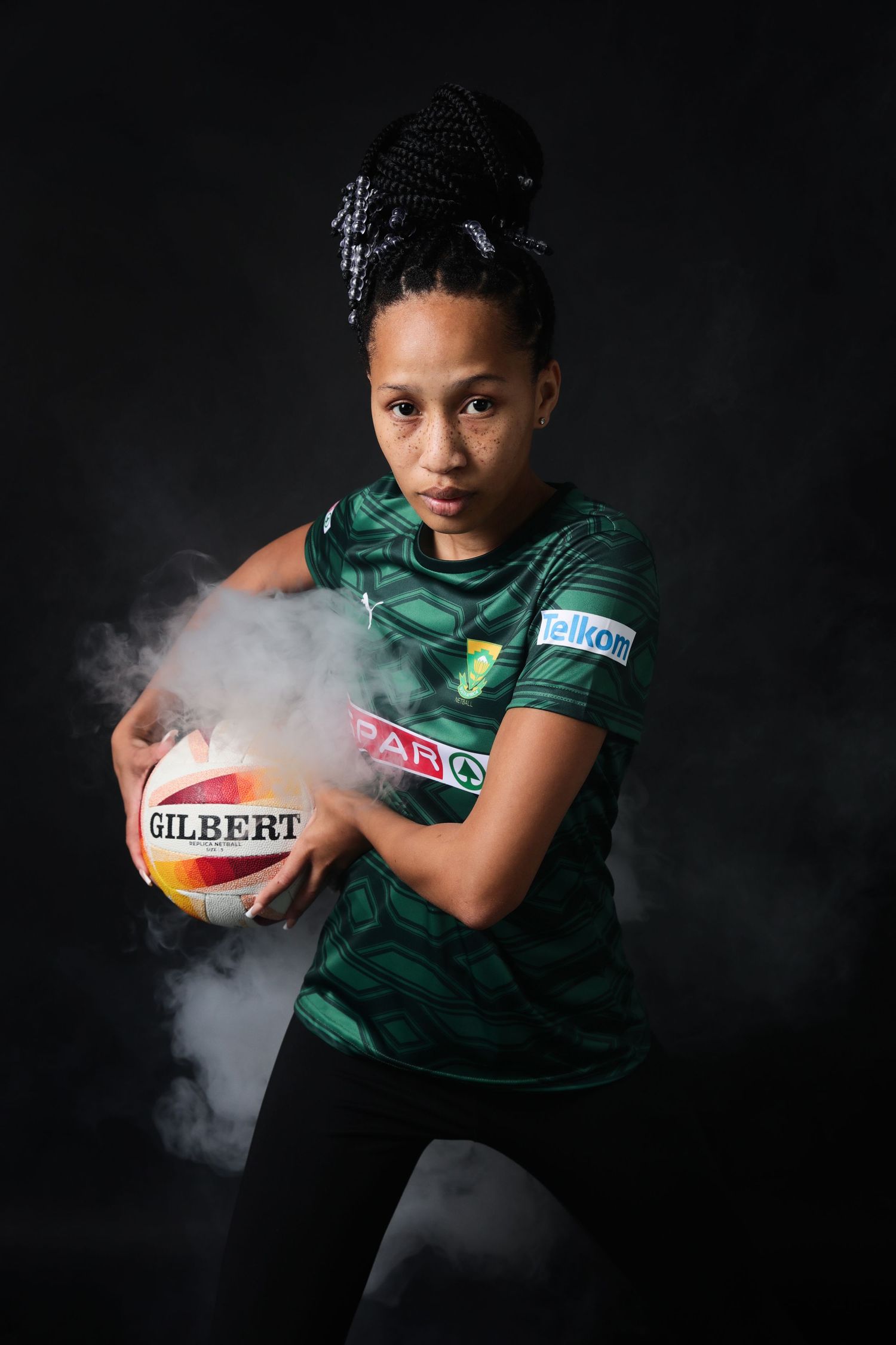 Female netball player in green Springboks SPAR jersey holds Gilbert ball with dramatic smoke effect.