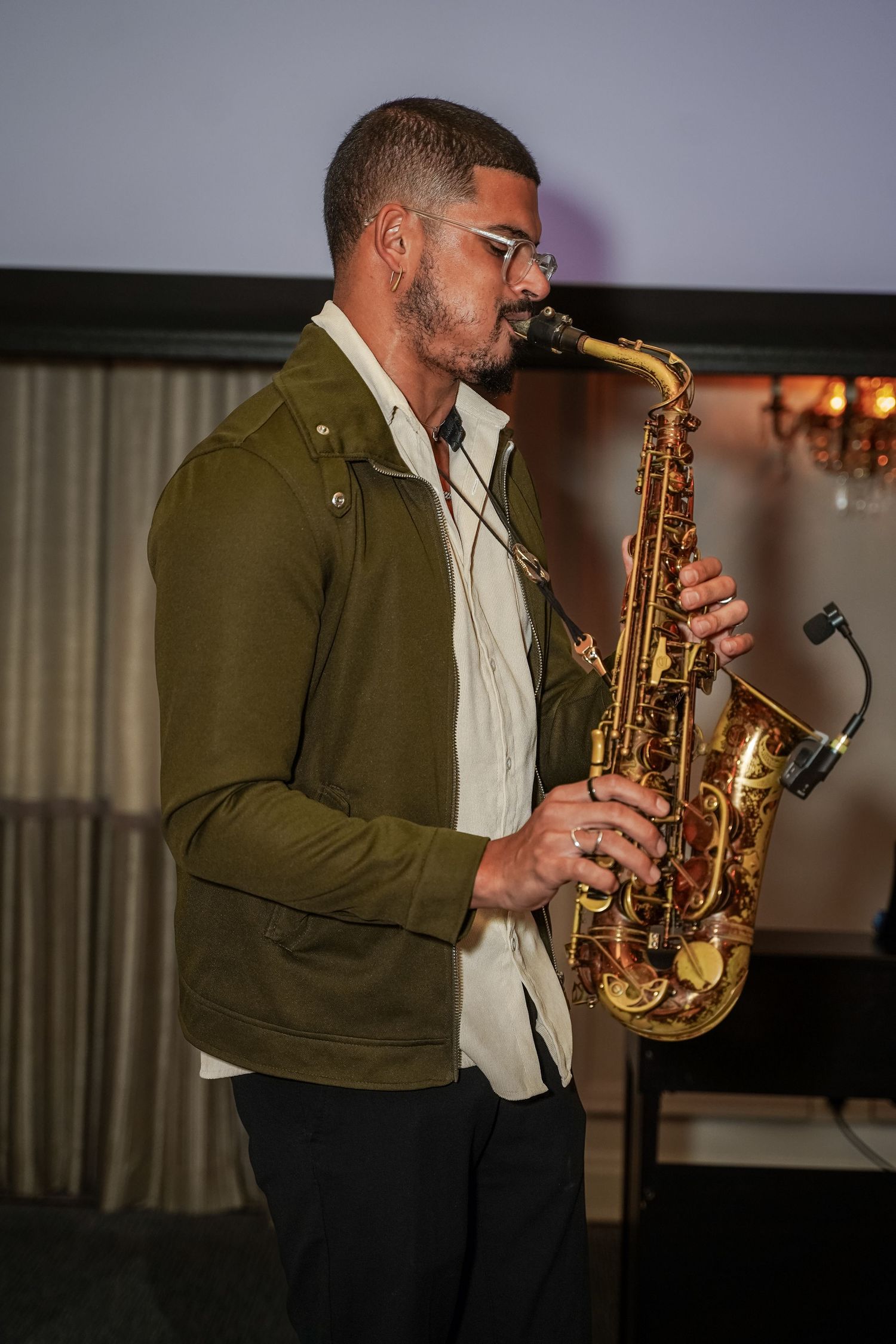 Musician in olive jacket playing a gold saxophone at an indoor event.