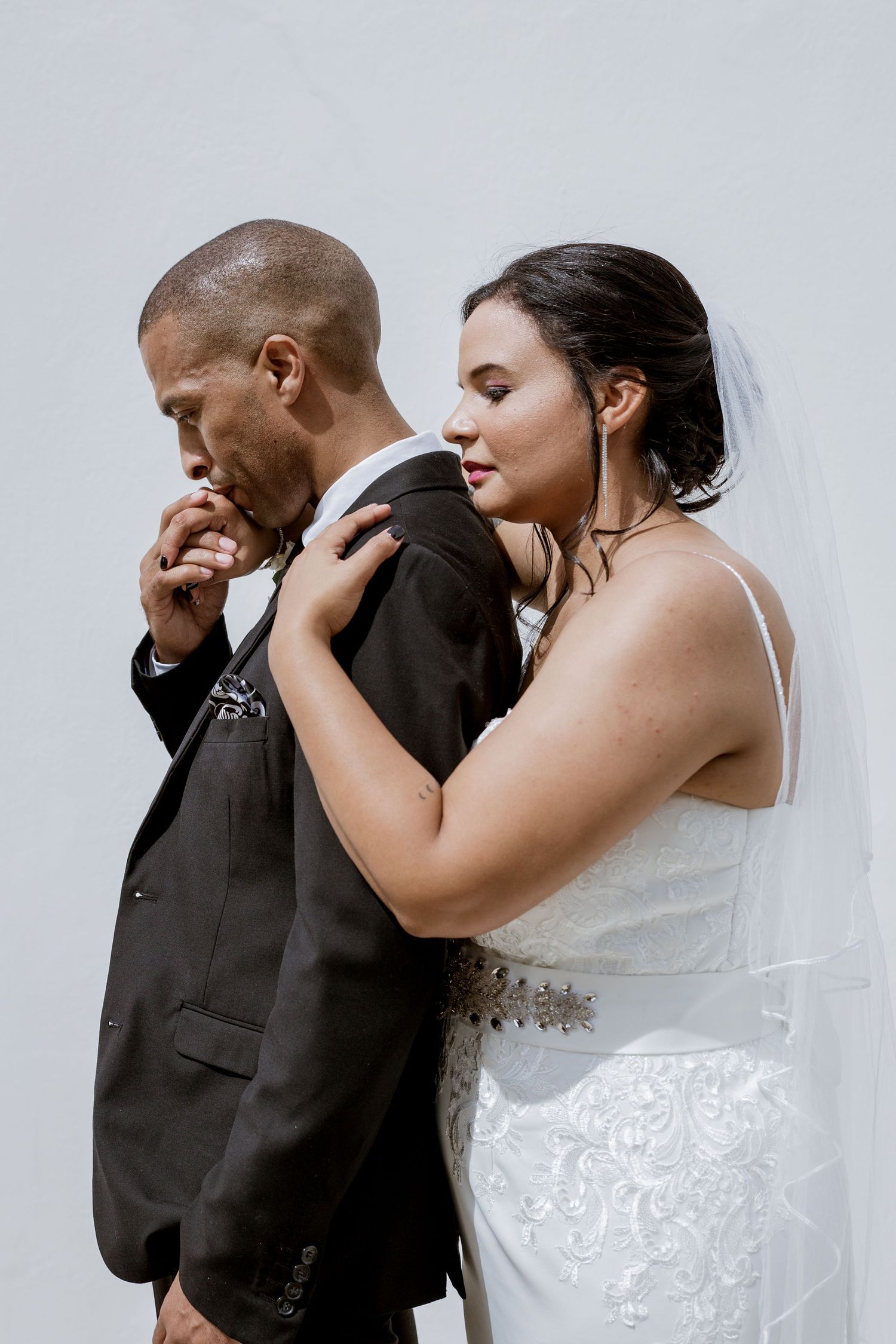 Groom in black suit kisses bride's hand as she embraces him, both in wedding attire against white background.