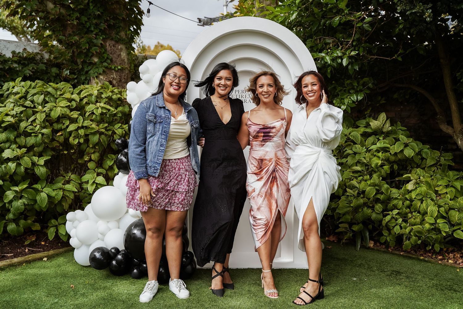Four women posing together in front of a white balloon arch outdoors.