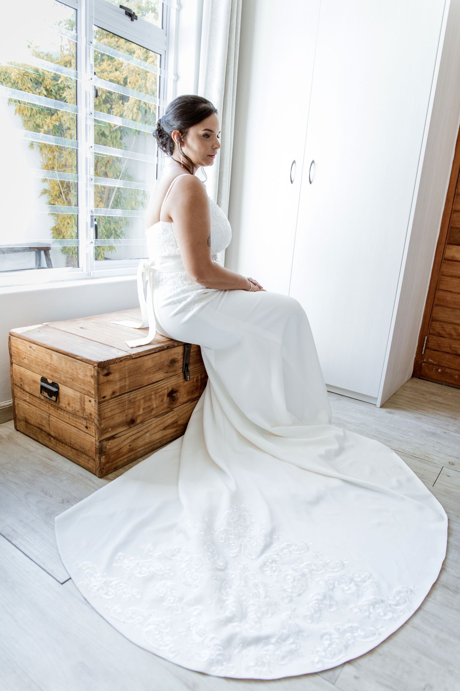 Bride in white satin wedding dress sitting on wooden chest by bright window.