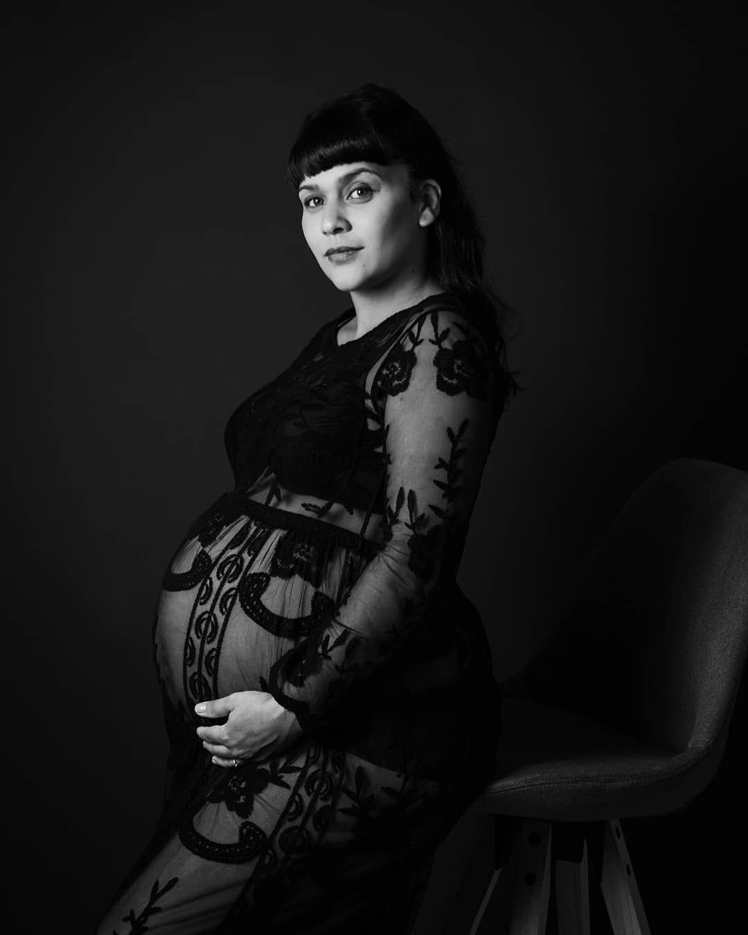 Pregnant woman in black lace maternity gown cradles baby bump in elegant black and white studio portrait.