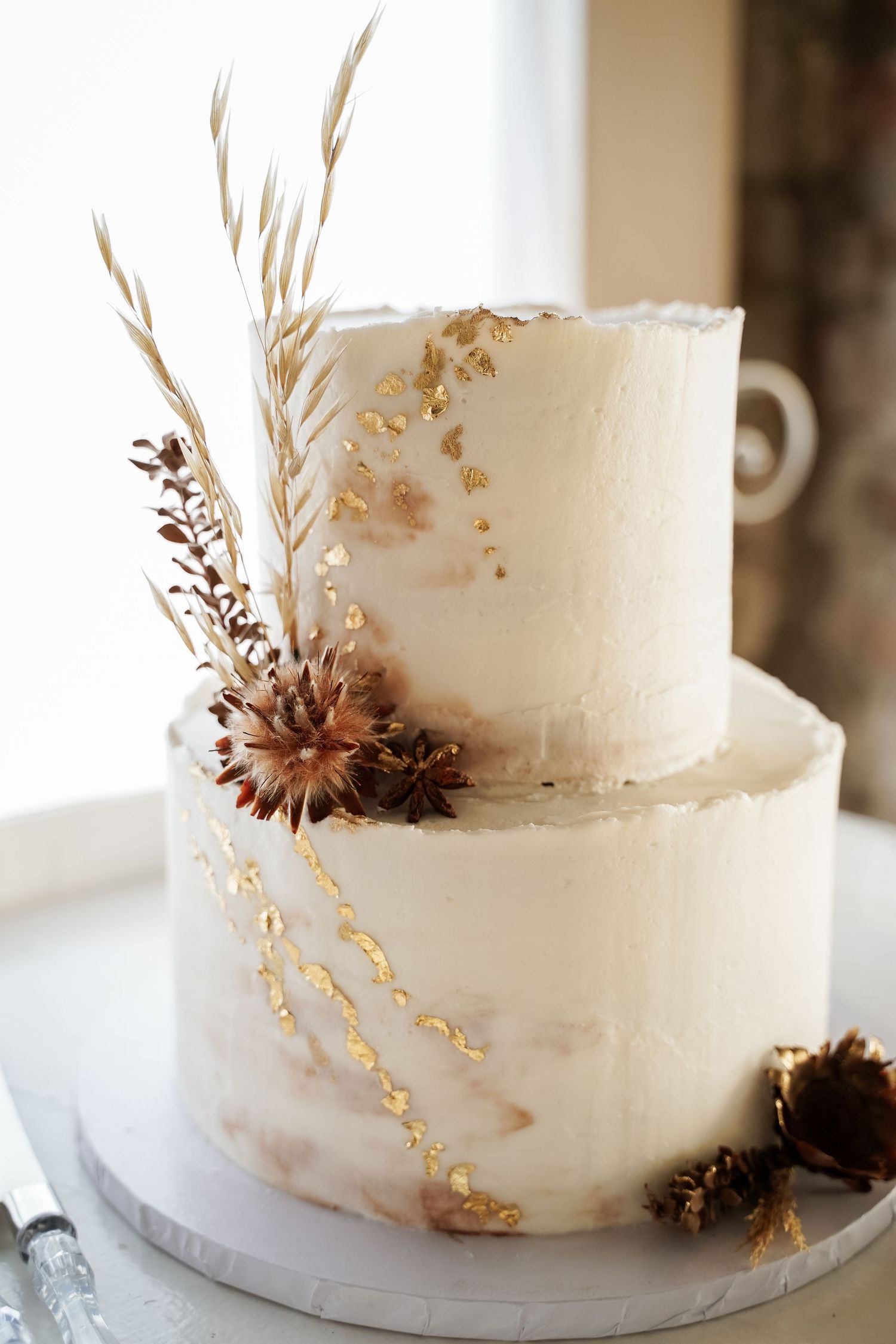 Elegant two-tier white wedding cake decorated with gold leaf, dried flowers, and wheat stalks.