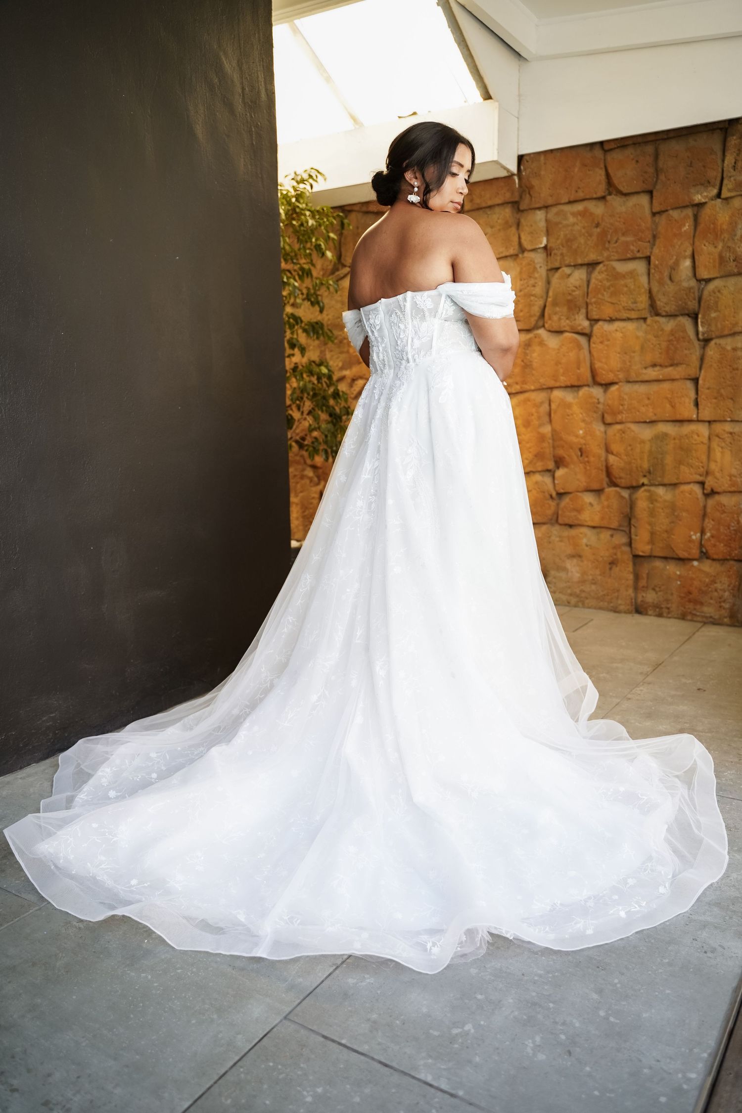 Bride in stunning off-shoulder white ball gown with long train viewed from behind near stone wall.