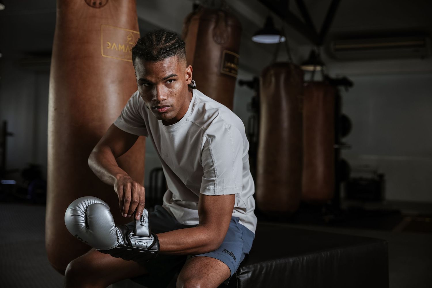 Young male boxer in white shirt rests with boxing gloves beside leather punching bags in a gym.