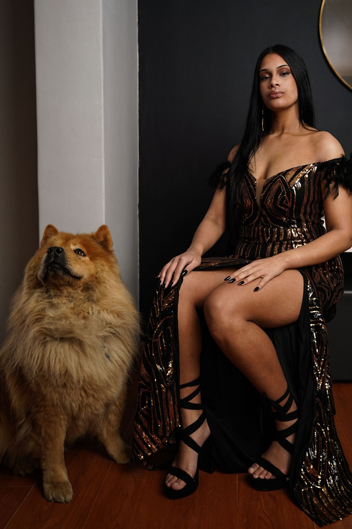 Woman in gold sequin gown seated beside a fluffy Chow Chow dog indoors.