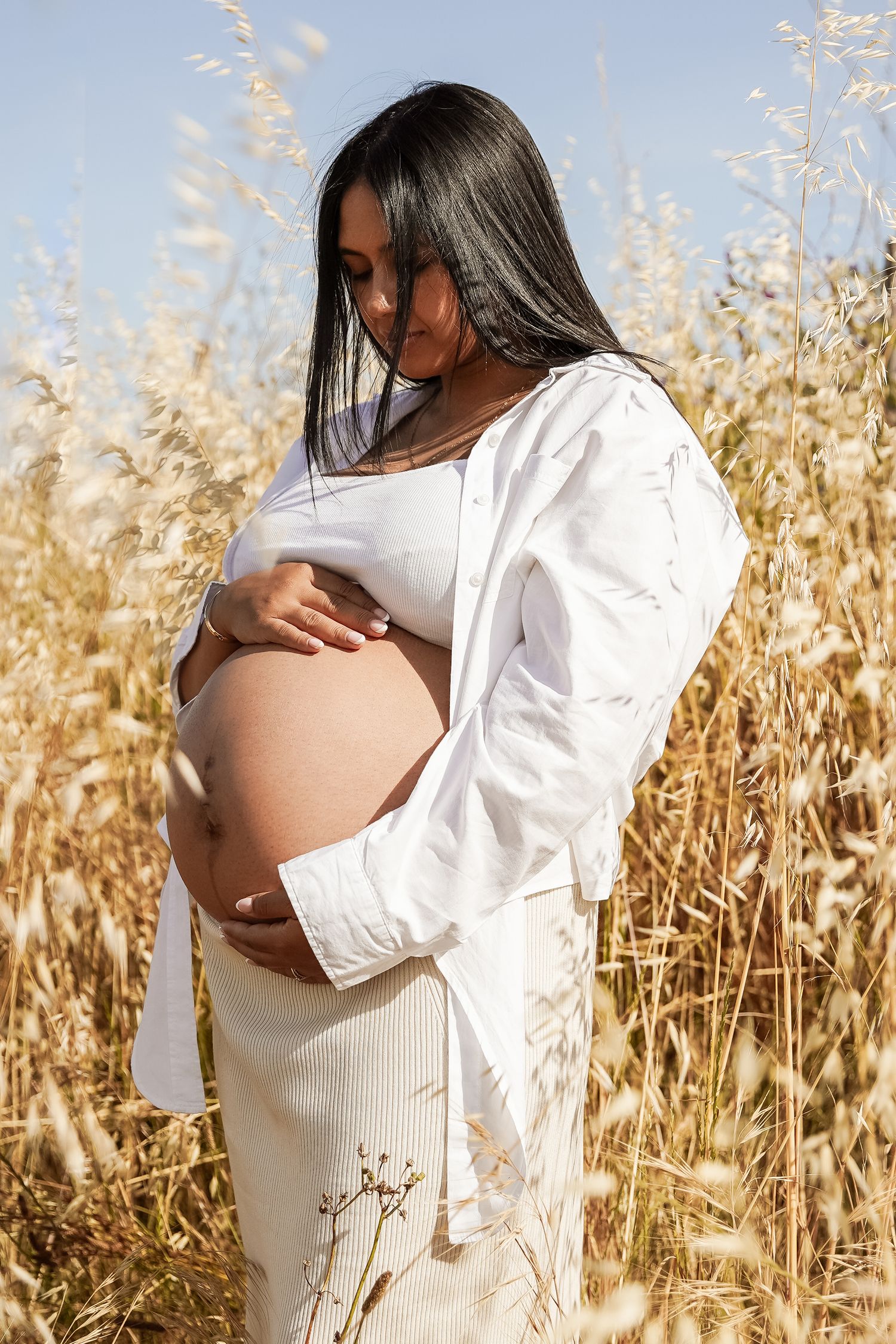 A pregnant woman in white clothing gently holds her bare bump while standing among tall golden grass in sunlight.