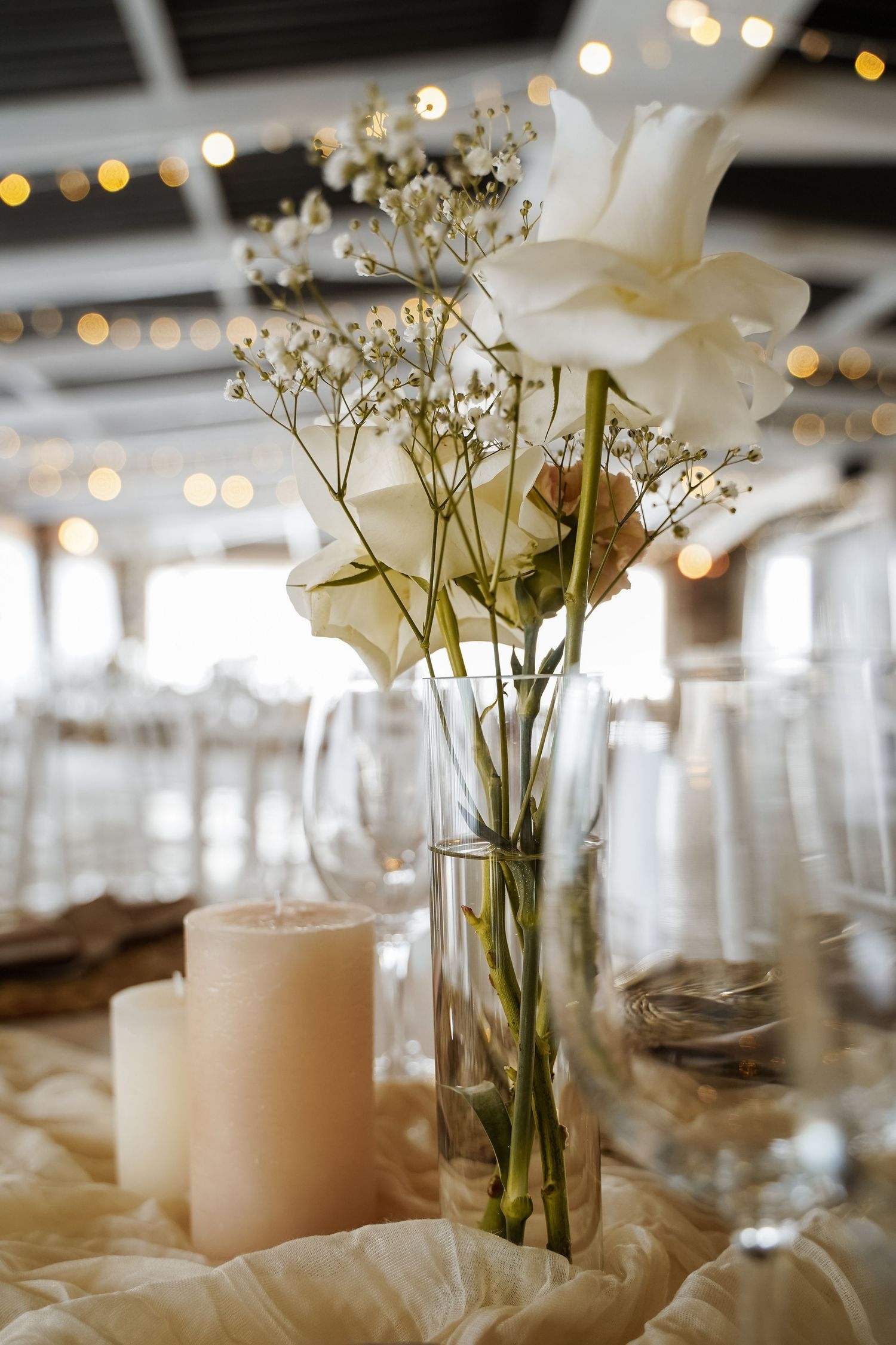Elegant wedding centerpiece with white roses, baby's breath in glass vase beside pillar candle.