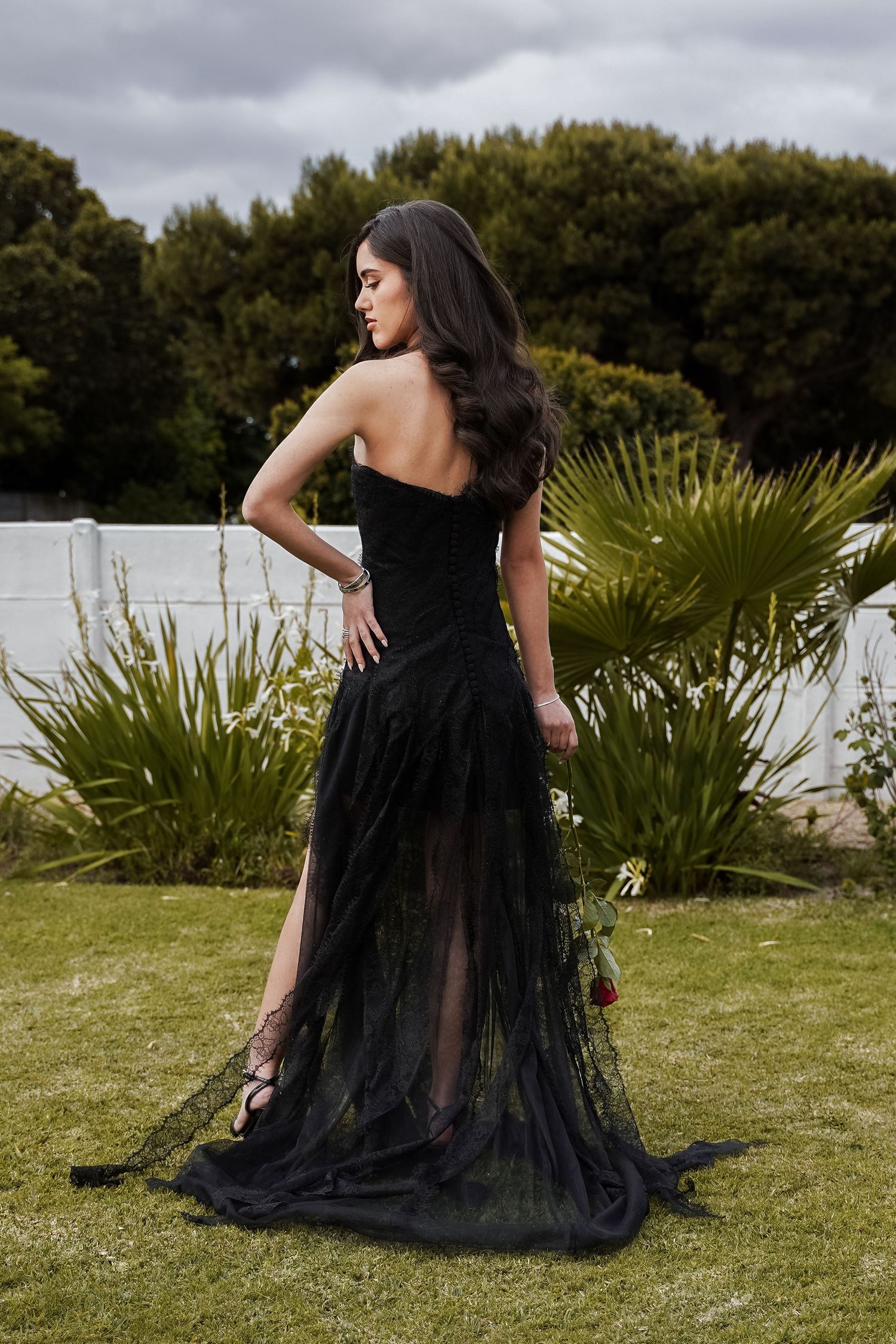 Young woman in black strapless tulle gown with high slit posing from behind in outdoor garden setting.