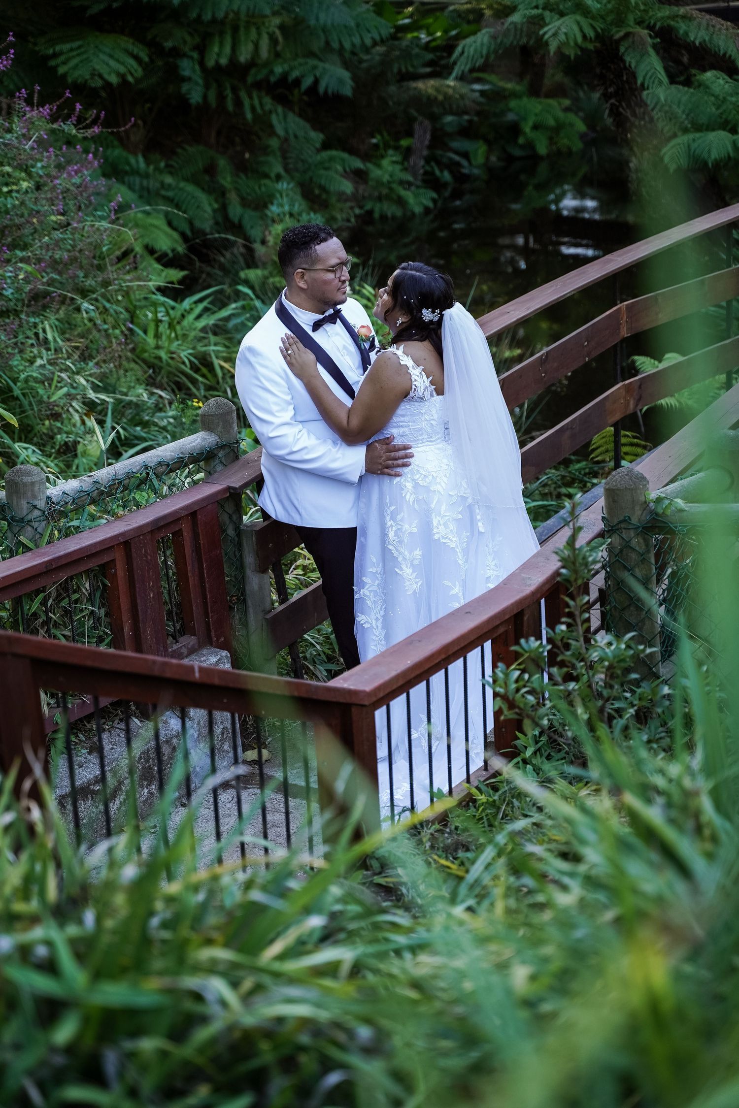 A newlywed couple in wedding attire embraces on a wooden bridge surrounded by lush green garden foliage.
