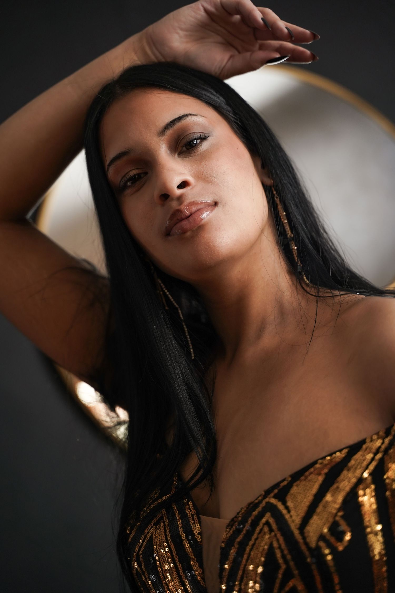 Close-up portrait of woman in gold sequin top posing by a round mirror.