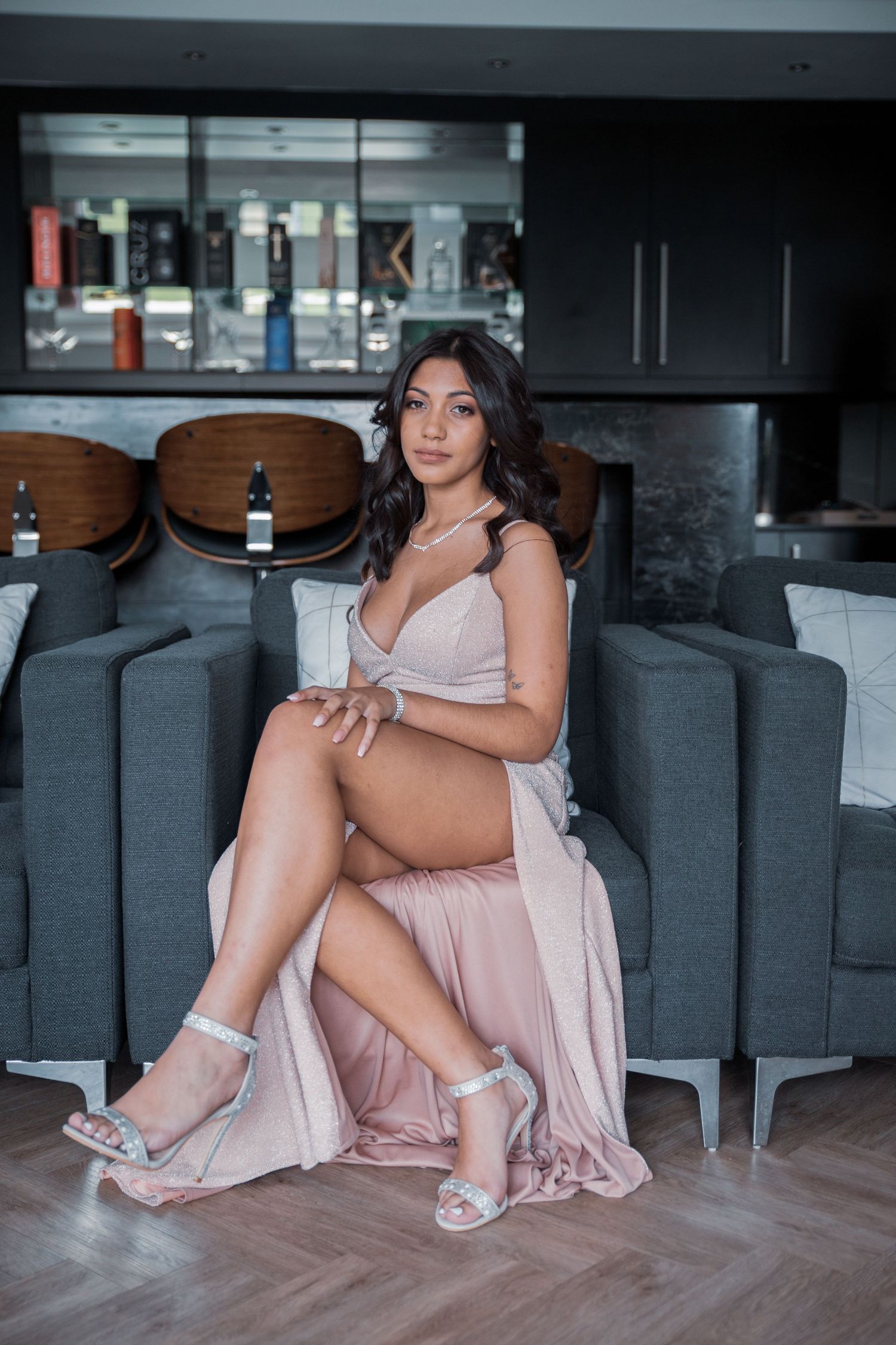 Woman in blush pink wrap dress with silver heels seated in modern lounge interior.