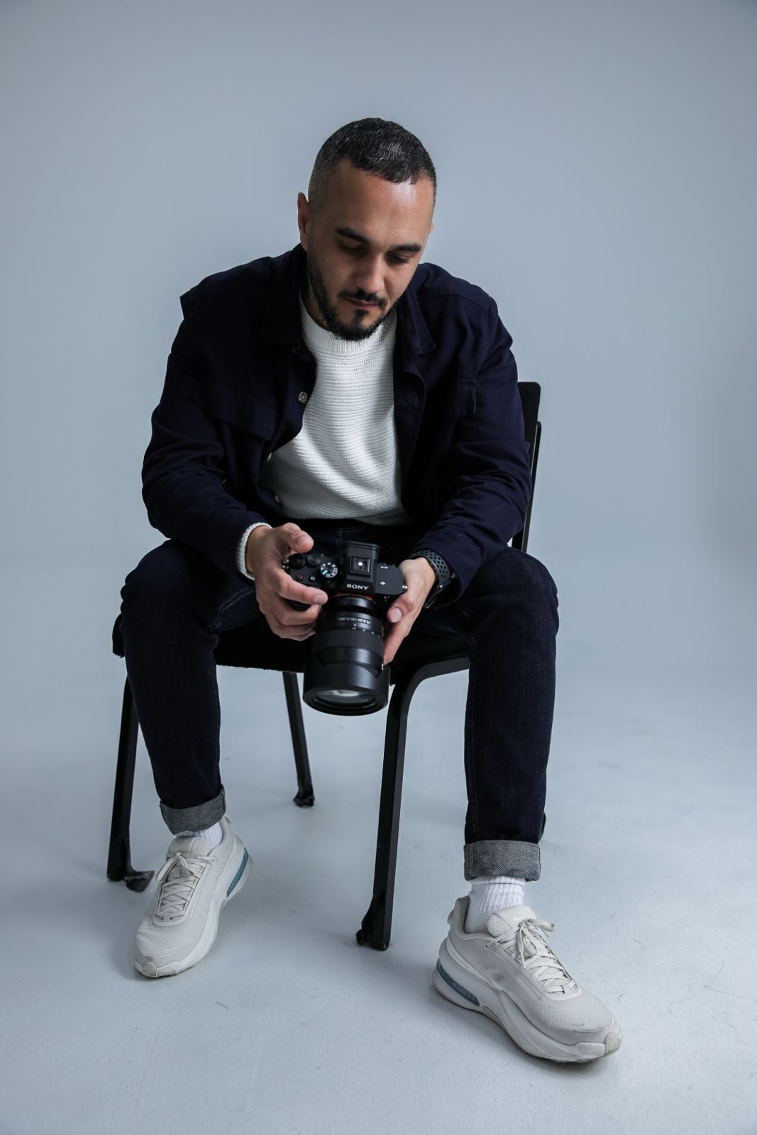 A male photographer sits on a chair holding a DSLR camera, wearing a dark jacket and white sneakers against a gray background.