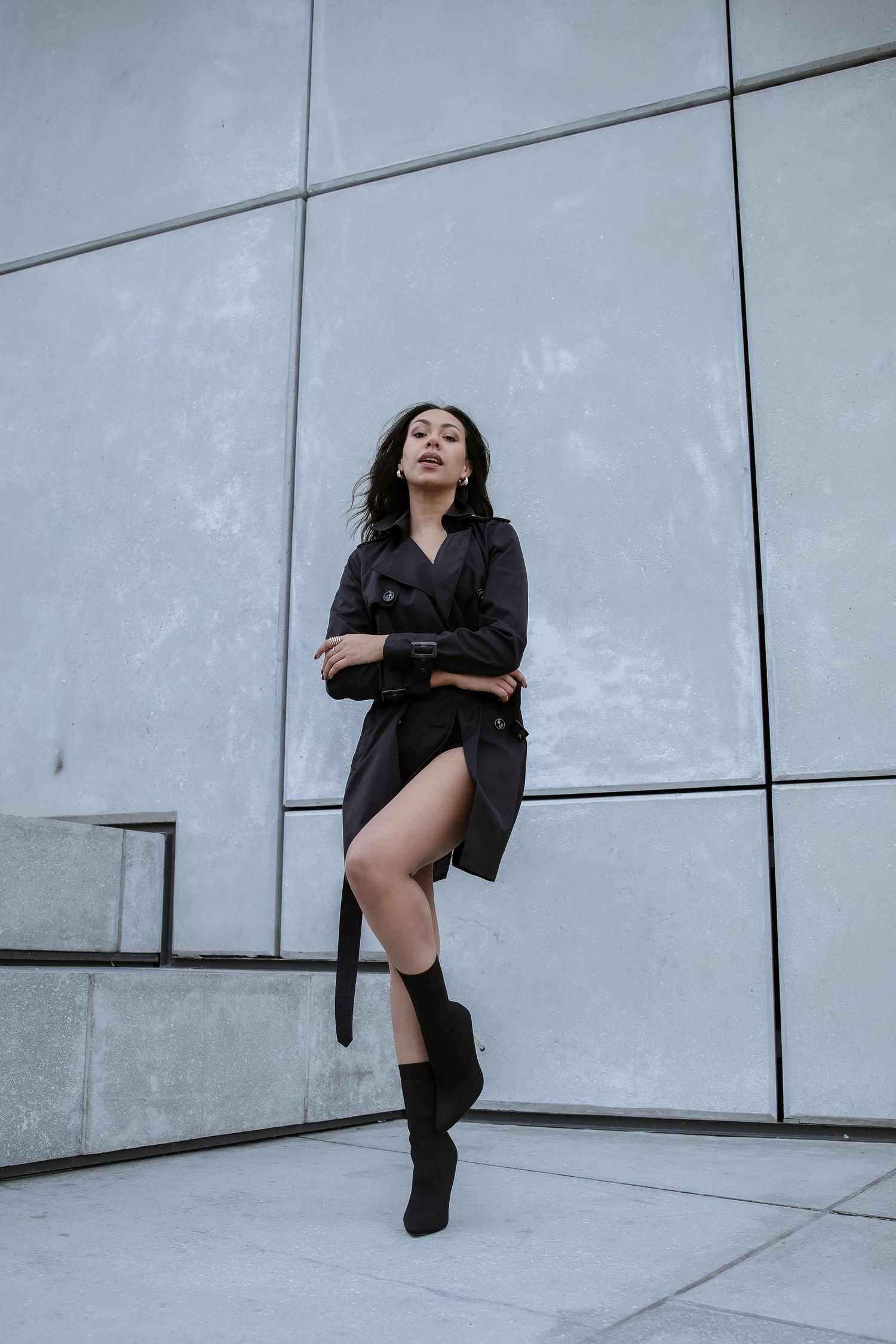 Stylish woman in black trench coat and boots posing confidently against modern gray wall.