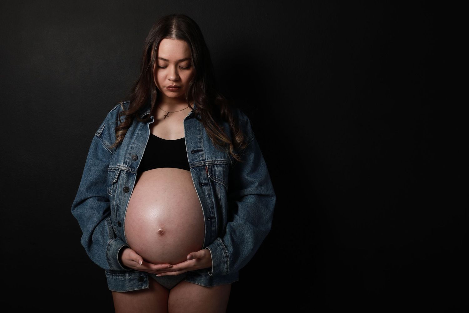 Pregnant woman in open denim jacket gazes down at bare baby bump on black background.