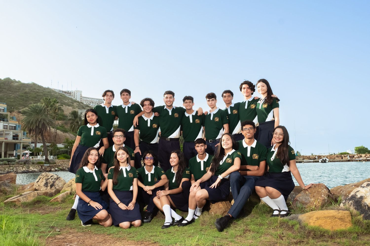 Students in green and navy uniforms pose for a group photo outdoors near a rocky waterfront on a sunny day.