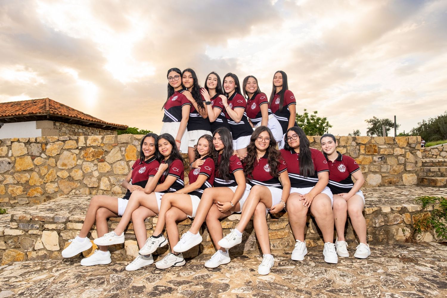 Group of teenage girls in matching red and white uniforms posing together on a stone wall outdoors.