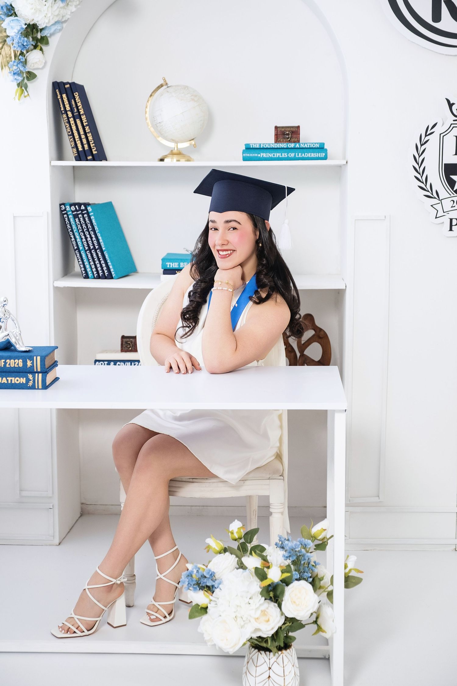 Smiling young woman in white dress and graduation cap sitting at white desk with bookshelf and flowers in bright studio.