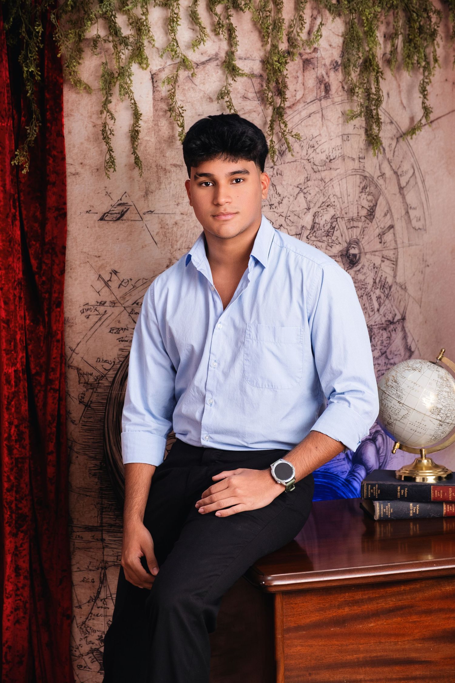 Young man in blue shirt and black pants sits on wooden desk with globe and books in decorative studio setting.