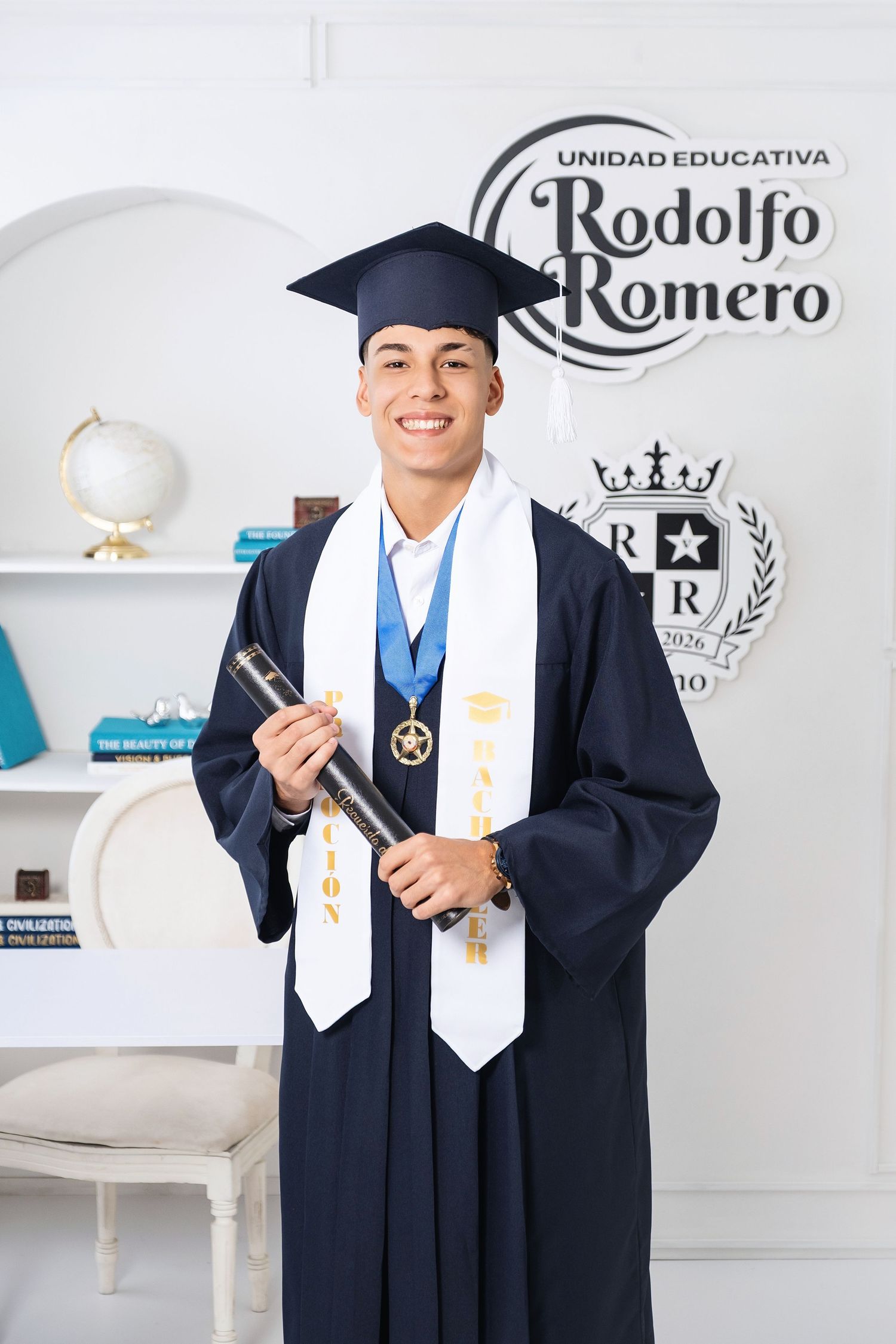 Smiling male graduate in cap and gown holds diploma at Unidad Educativa Rodolfo Romero.