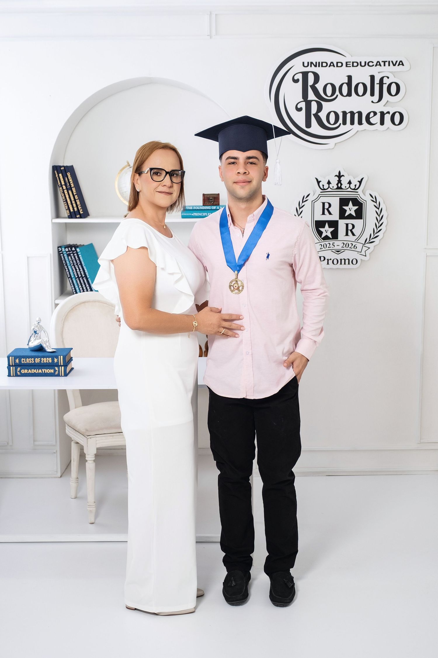 Graduate in cap and medal poses with woman in white dress at Unidad Educativa Rodolfo Romero graduation ceremony.