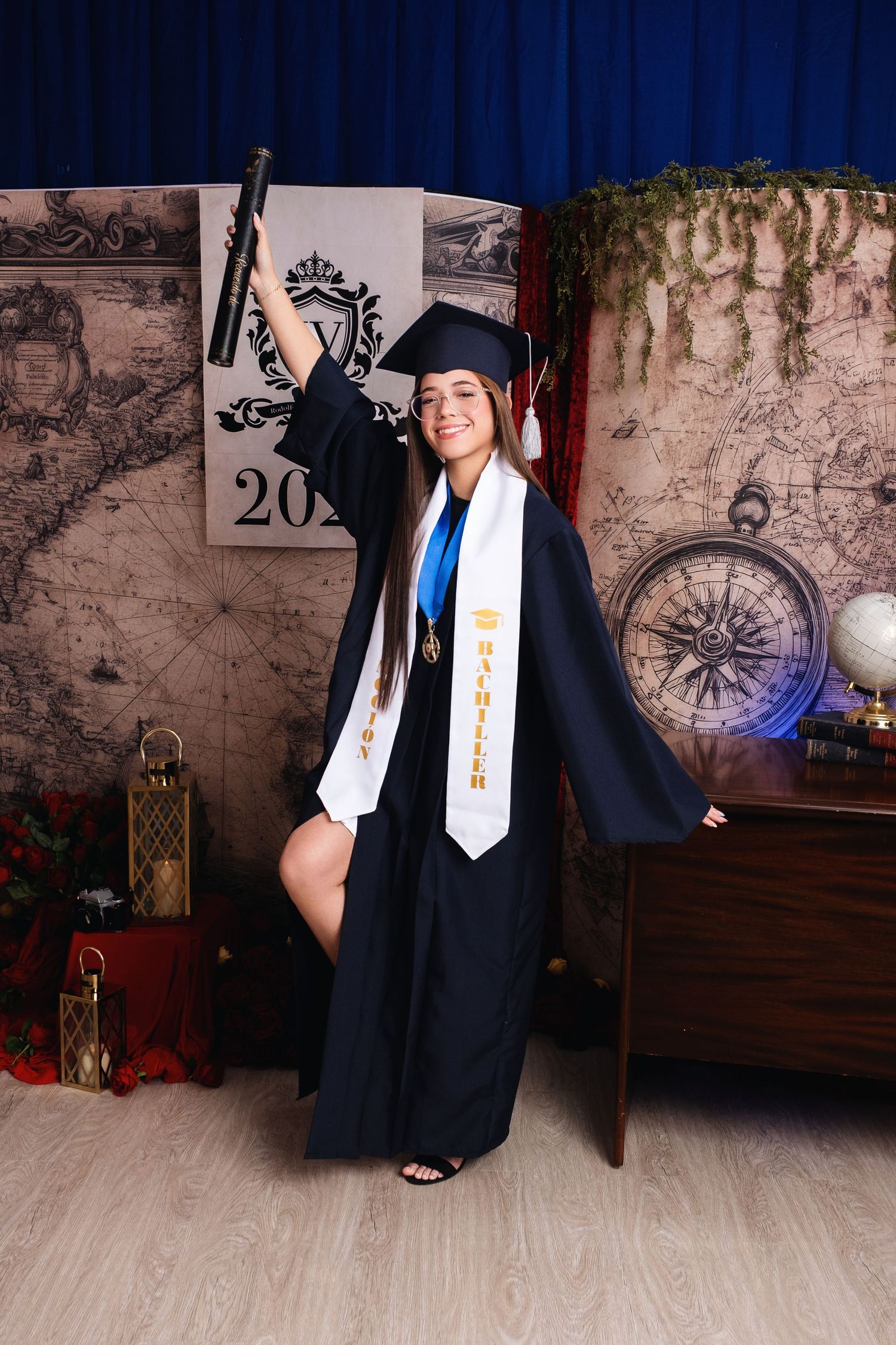 Young woman in black graduation cap and gown holding diploma, wearing Bachelor sash, posing in decorated photo studio.