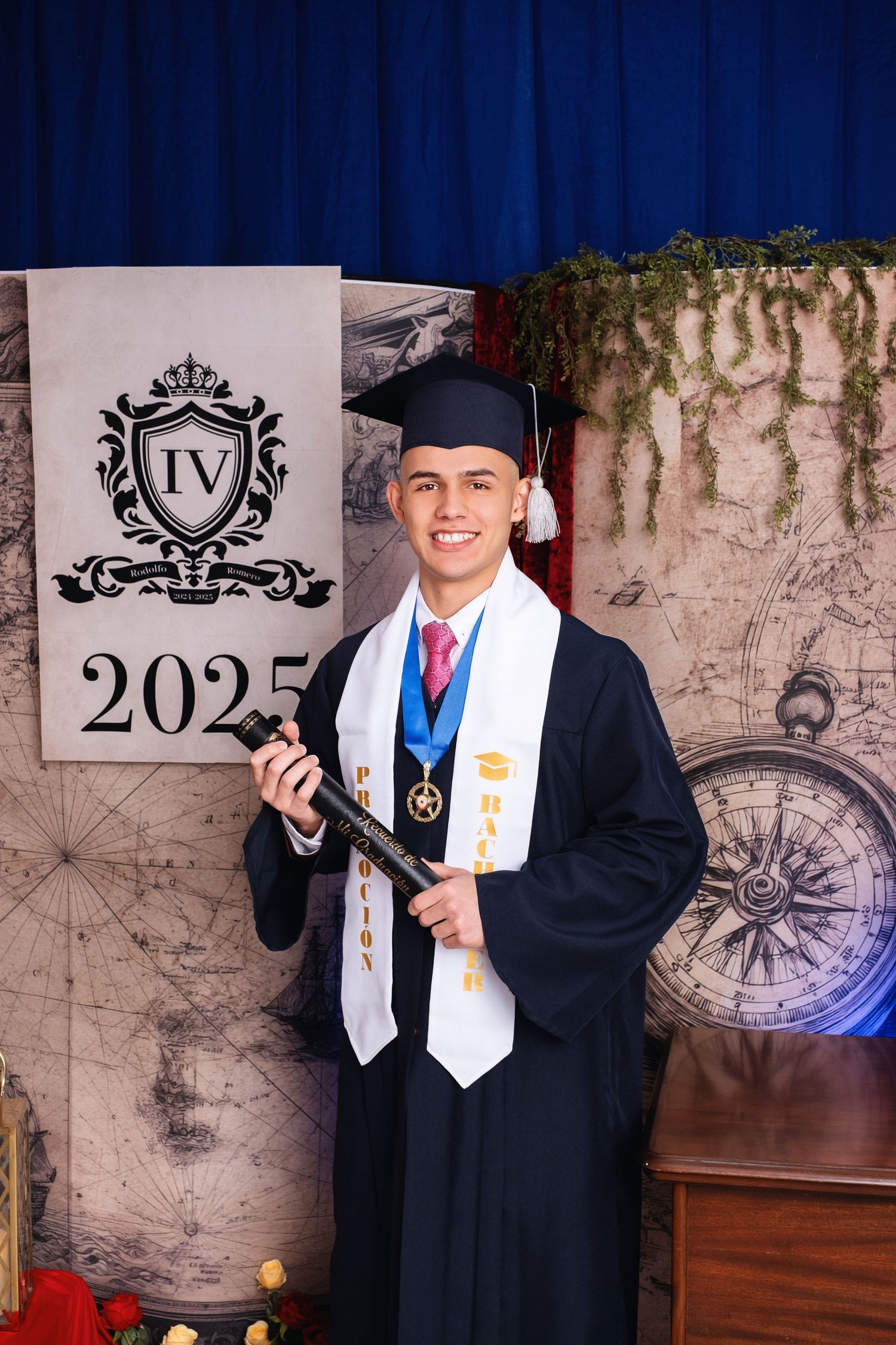Smiling graduate in cap and gown holds diploma, wearing medals, posing before a 2025 graduation backdrop.