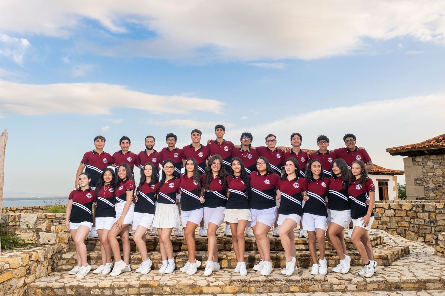 Large group of young athletes in matching maroon and white uniforms posing together outdoors on a stone terrace.