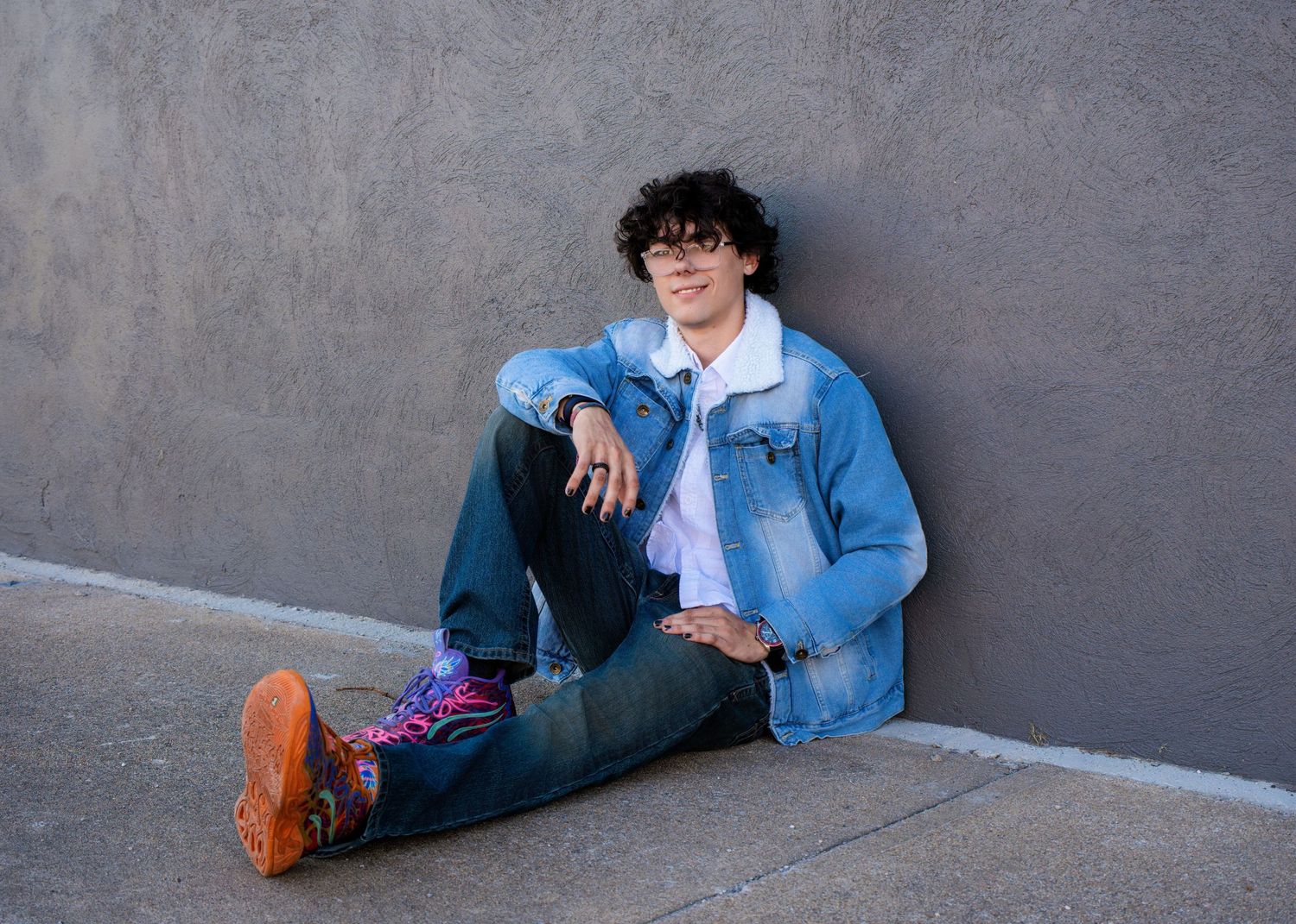 Young man in denim jacket and jeans sitting against gray wall wearing colorful socks and orange shoes.