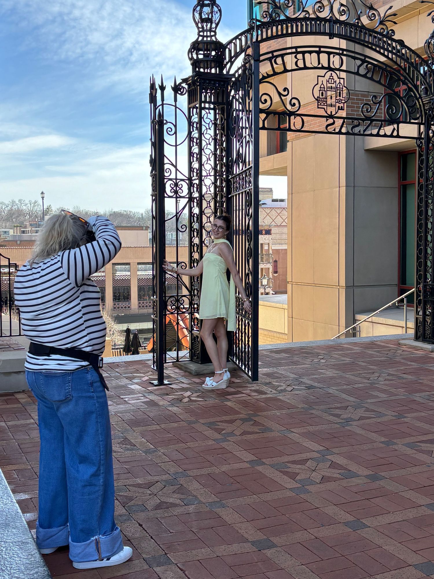 Two people stand on a terrace with ornate wrought iron gates and red tile flooring overlooking a city view.
