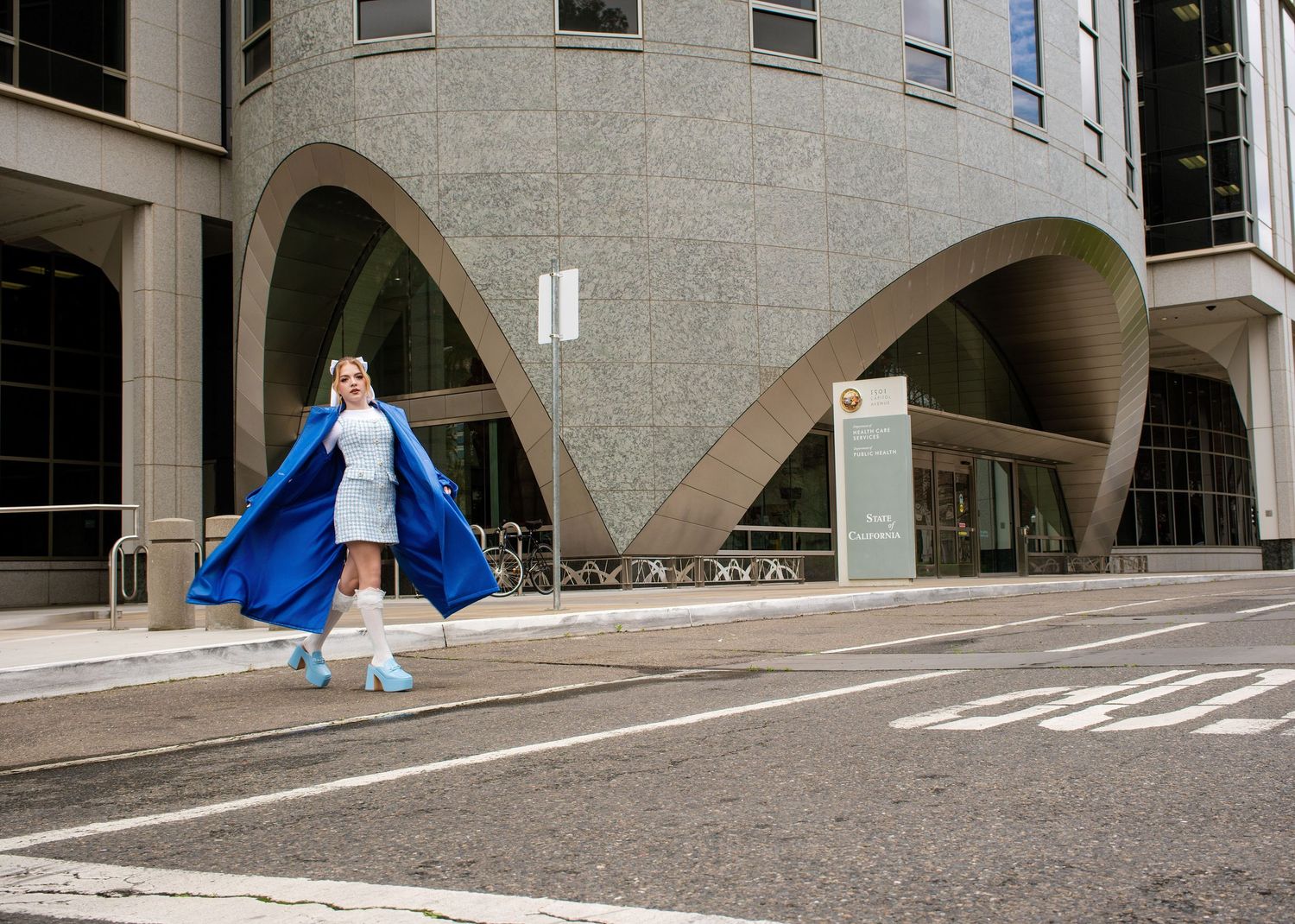 A figure in a flowing blue cape walks confidently past modern curved architecture on an urban street.