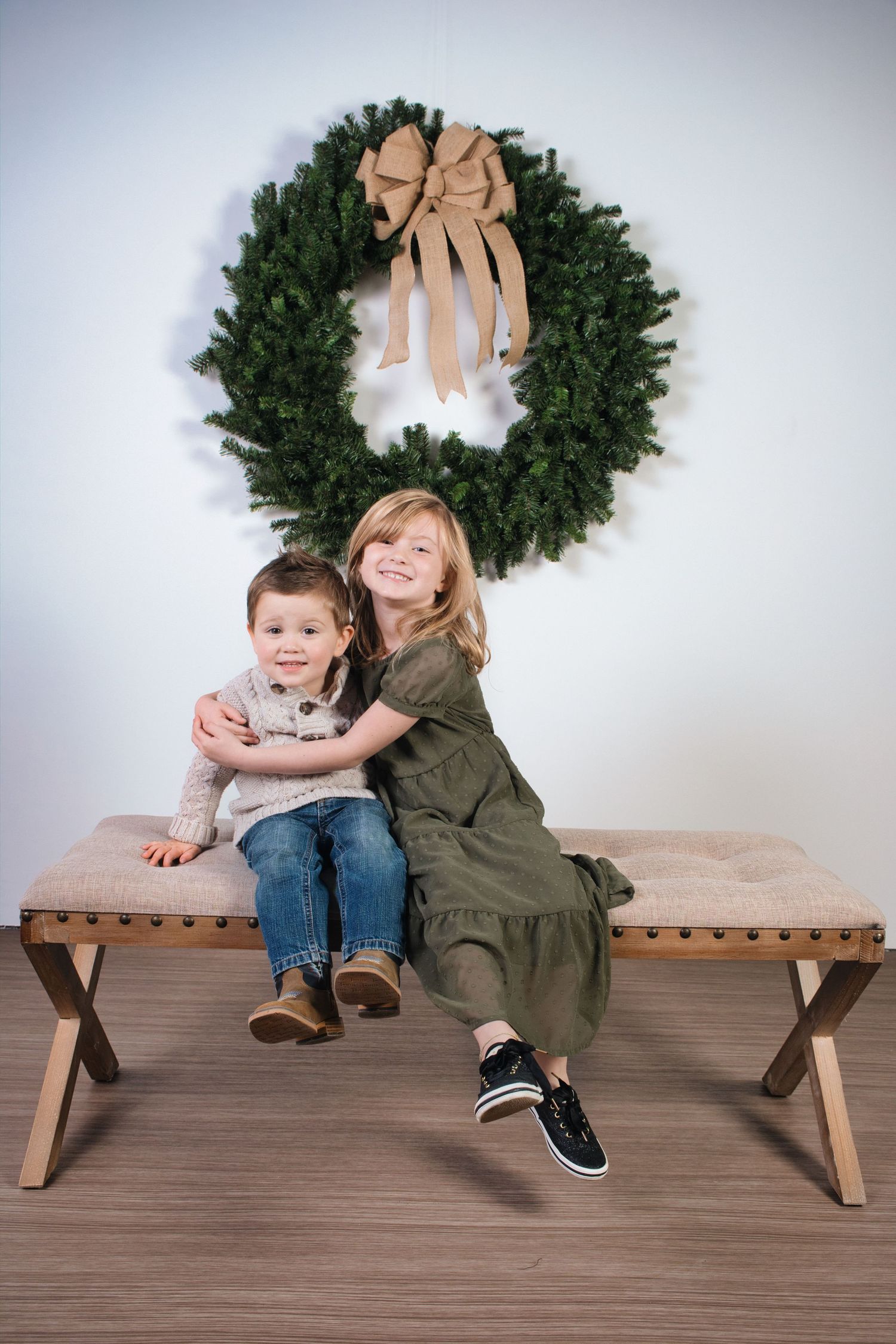 Two children sit together on wooden bench under large Christmas wreath with tan bow.