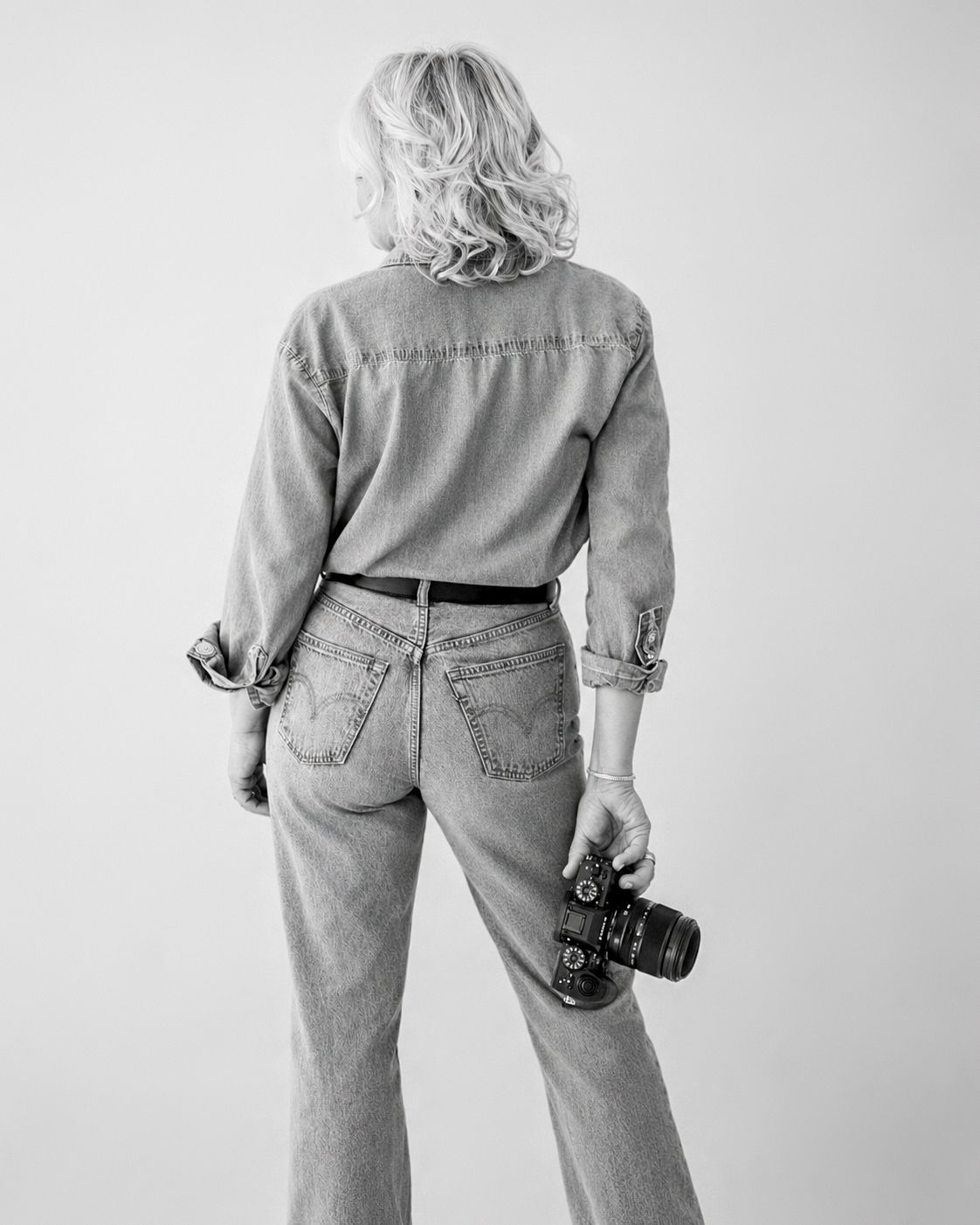 Black and white photo of a blonde woman from behind, wearing denim outfit, holding a camera.