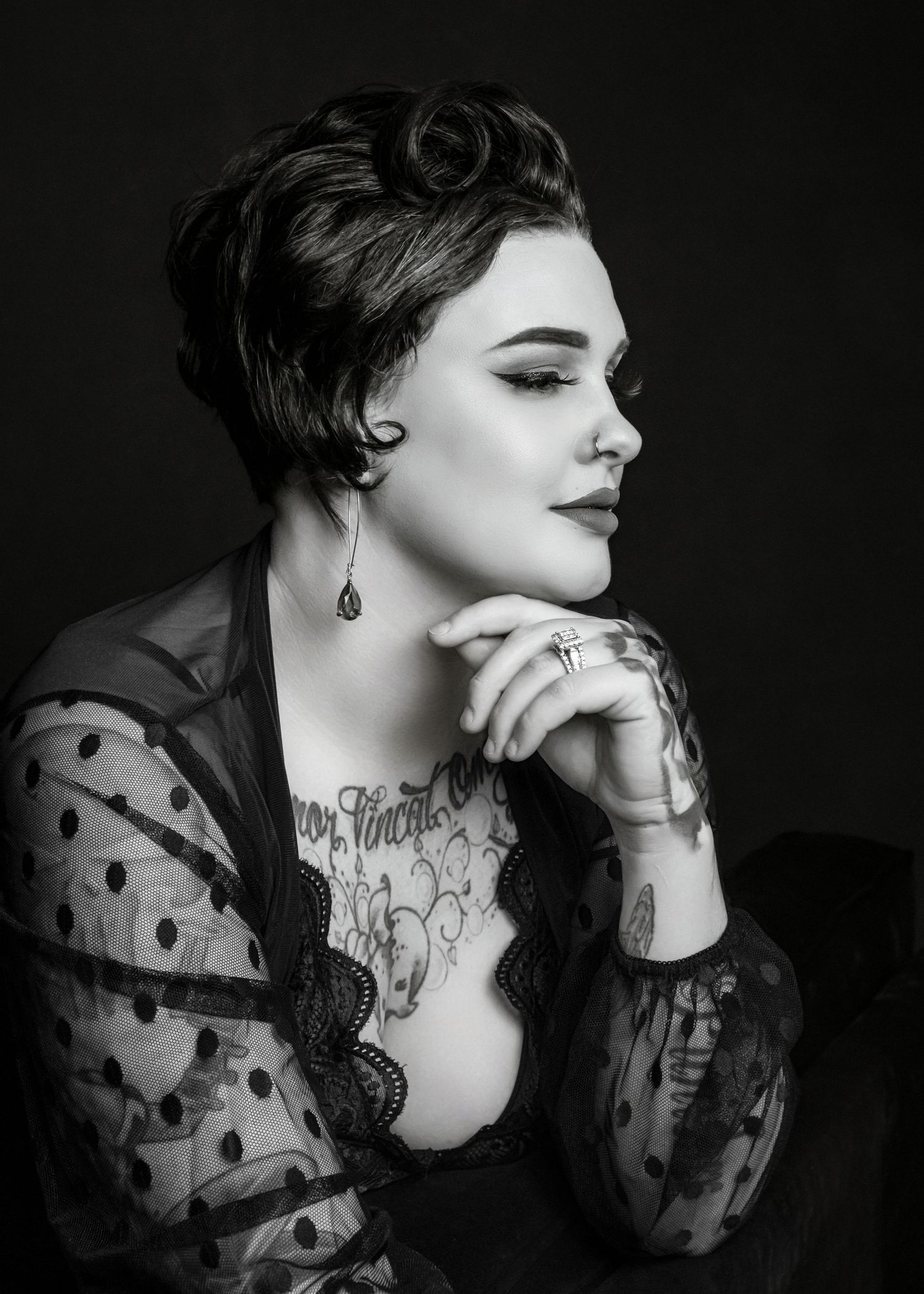 Black and white portrait of a tattooed woman in polka dot sheer top, elegant updo, dangling earring, hands clasped.